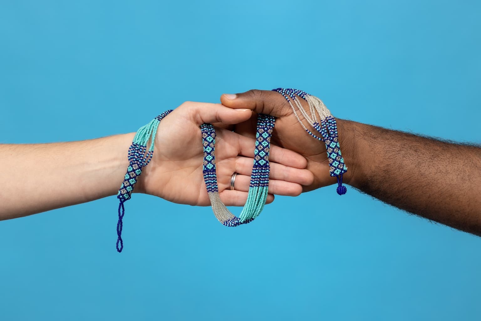 Two hands, one lighter and one darker, joined by a blue and turquoise beaded necklace against a bright blue background